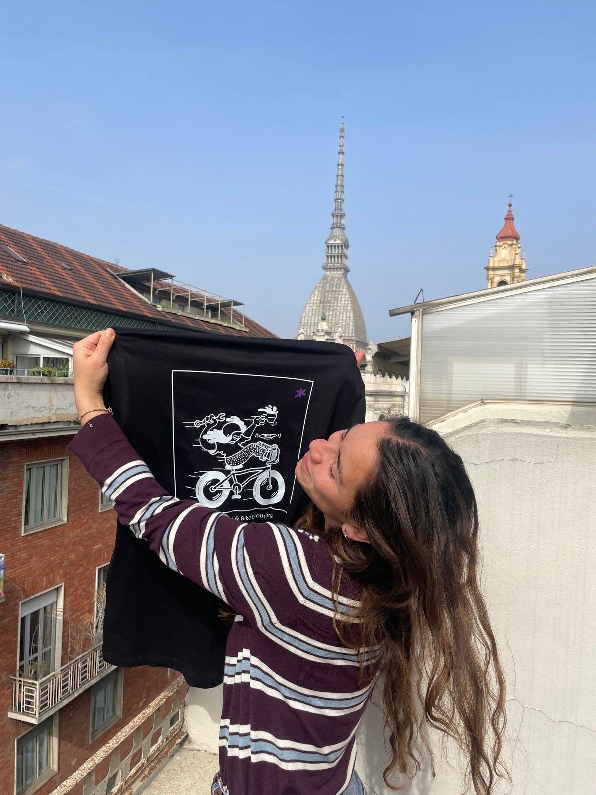 A woman on a Turin rooftop holding up a black Asterisco t-shirt, the Mole Antonelliana behind her