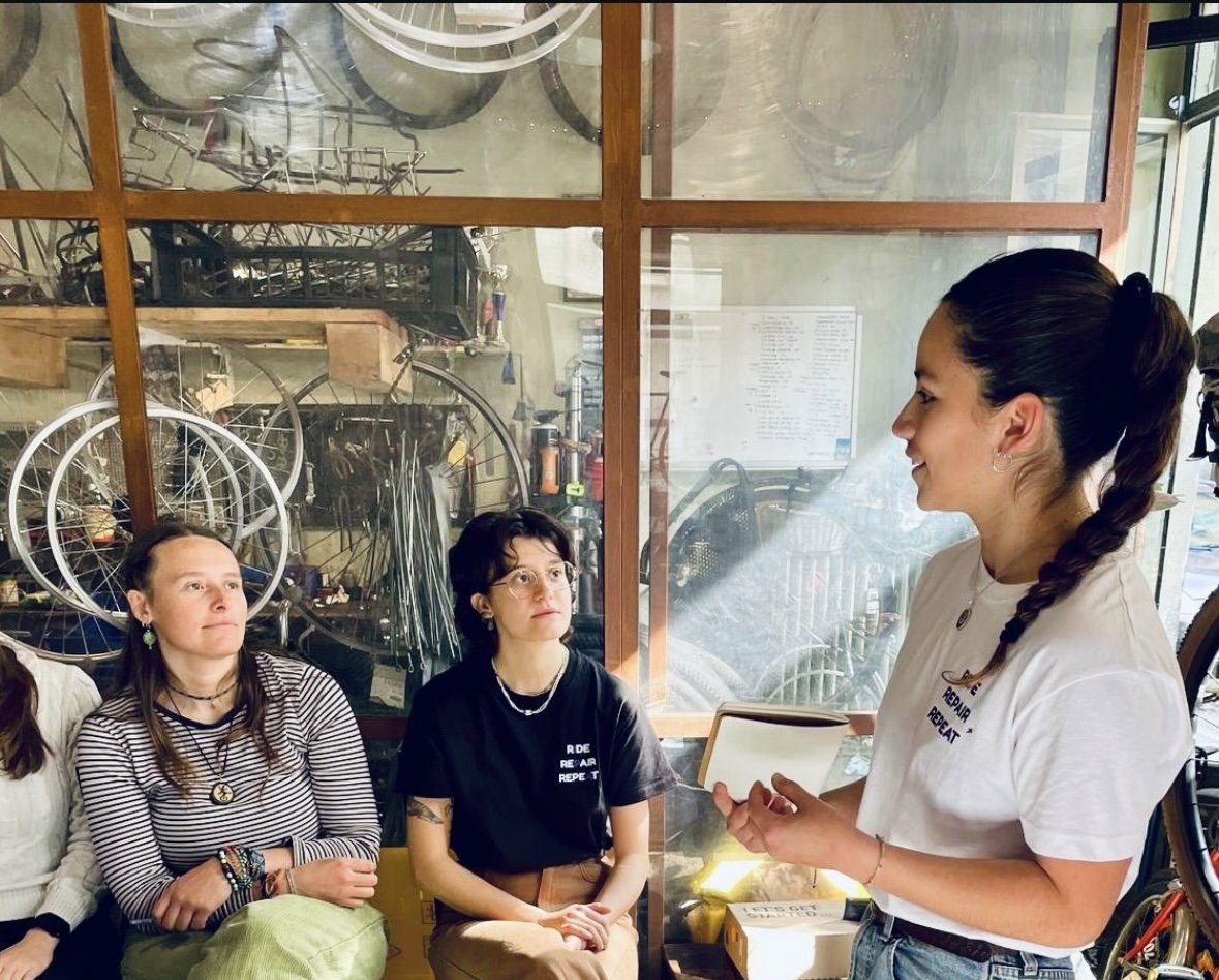 A woman demonstrating bicycle repair to two other women sitting and watching in a Turin workshop