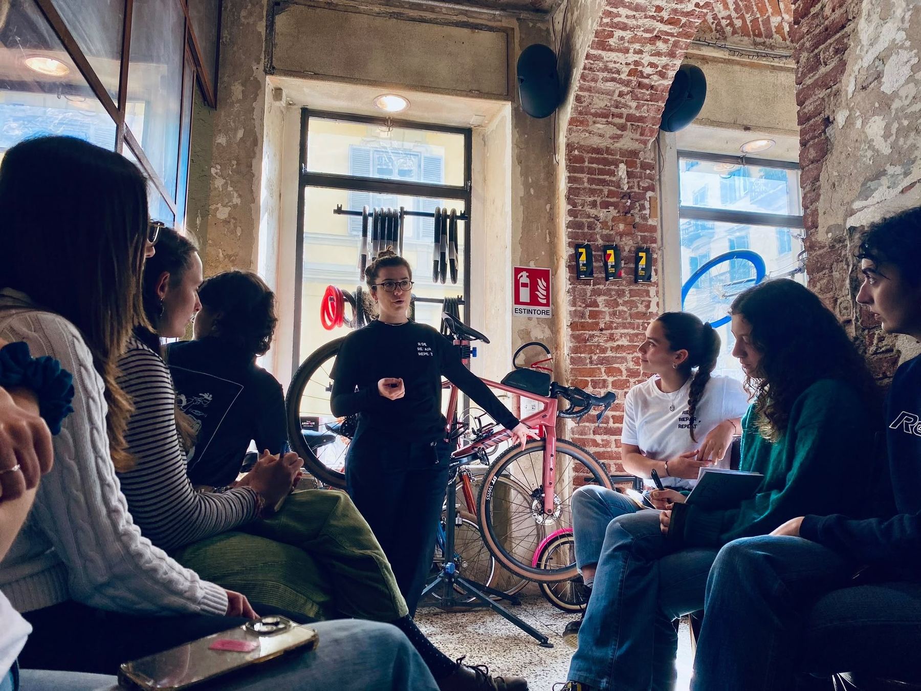 Women sitting in a circle in a Turin bike shop, listening to another woman standing beside a bicycle
