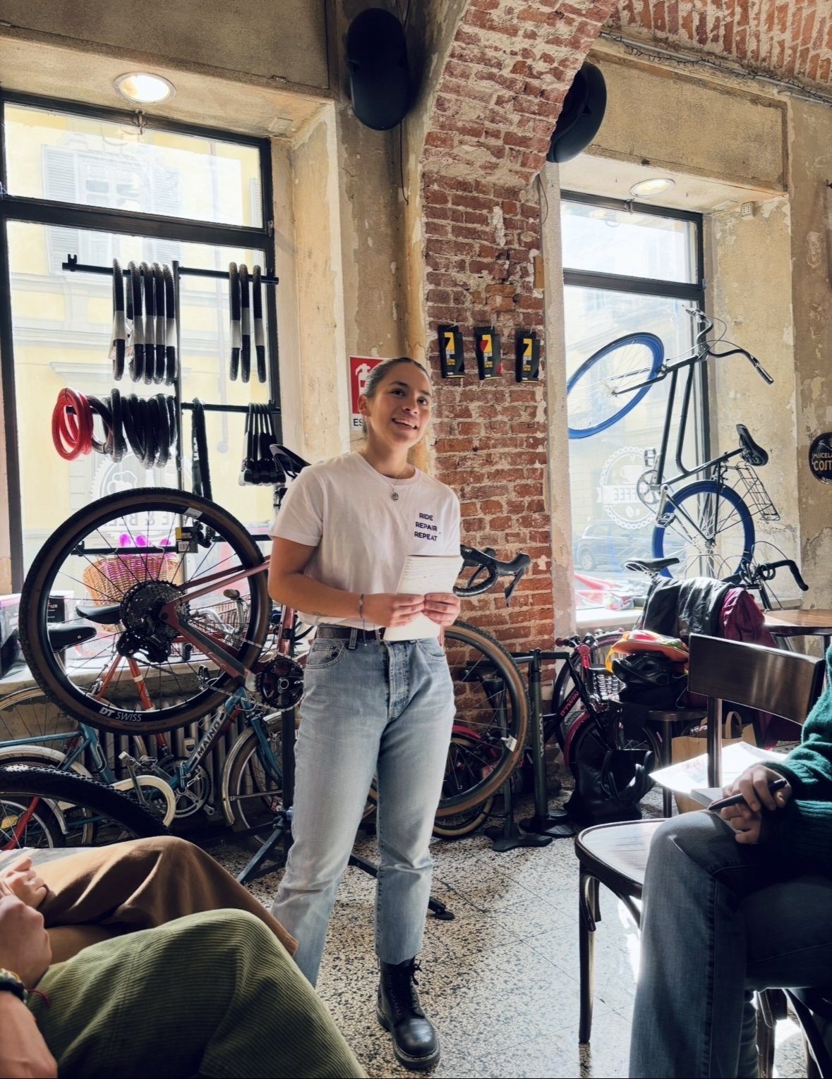 A woman standing in a bike workshop in Turin, holding notes and speaking to a seated group
