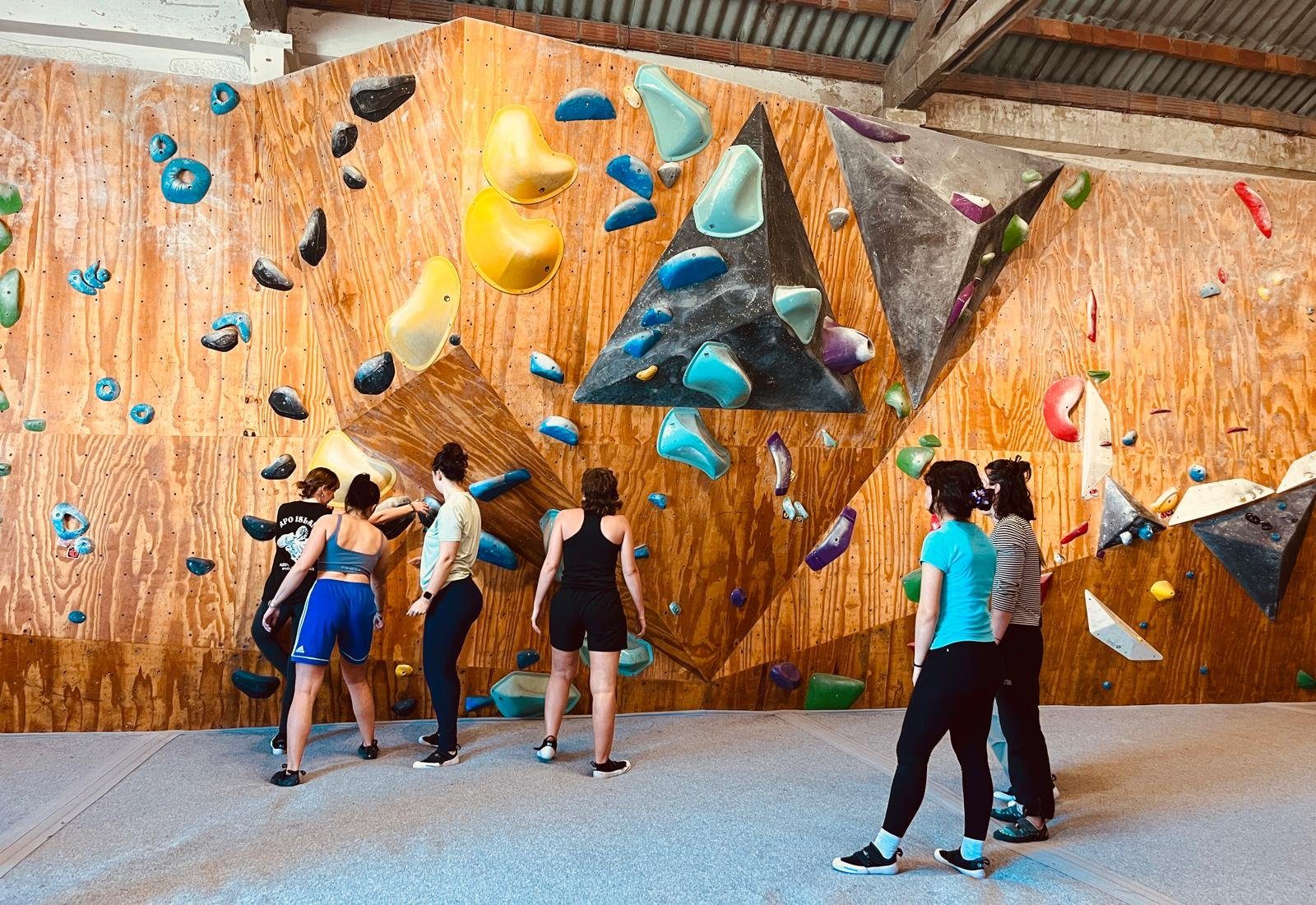 Six women standing at a tall indoor bouldering wall in Porto, working out a route together