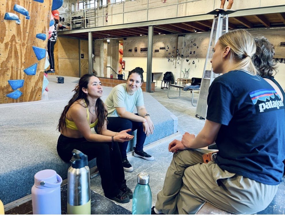 Three women sitting and talking on crash mats at a climbing gym in Porto