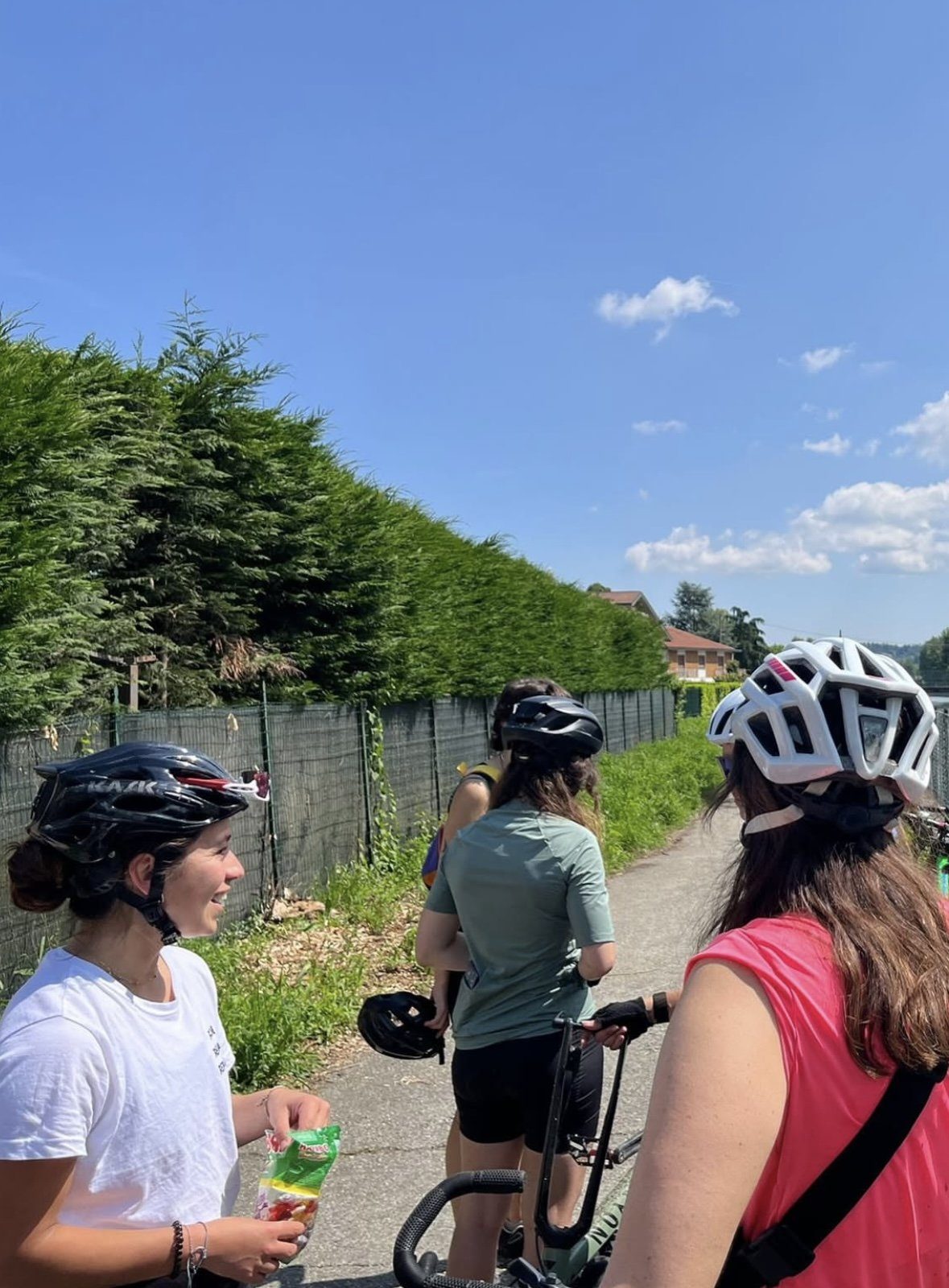 Three women in cycling helmets pausing on a sunny path outside Turin, chatting and sharing sweets