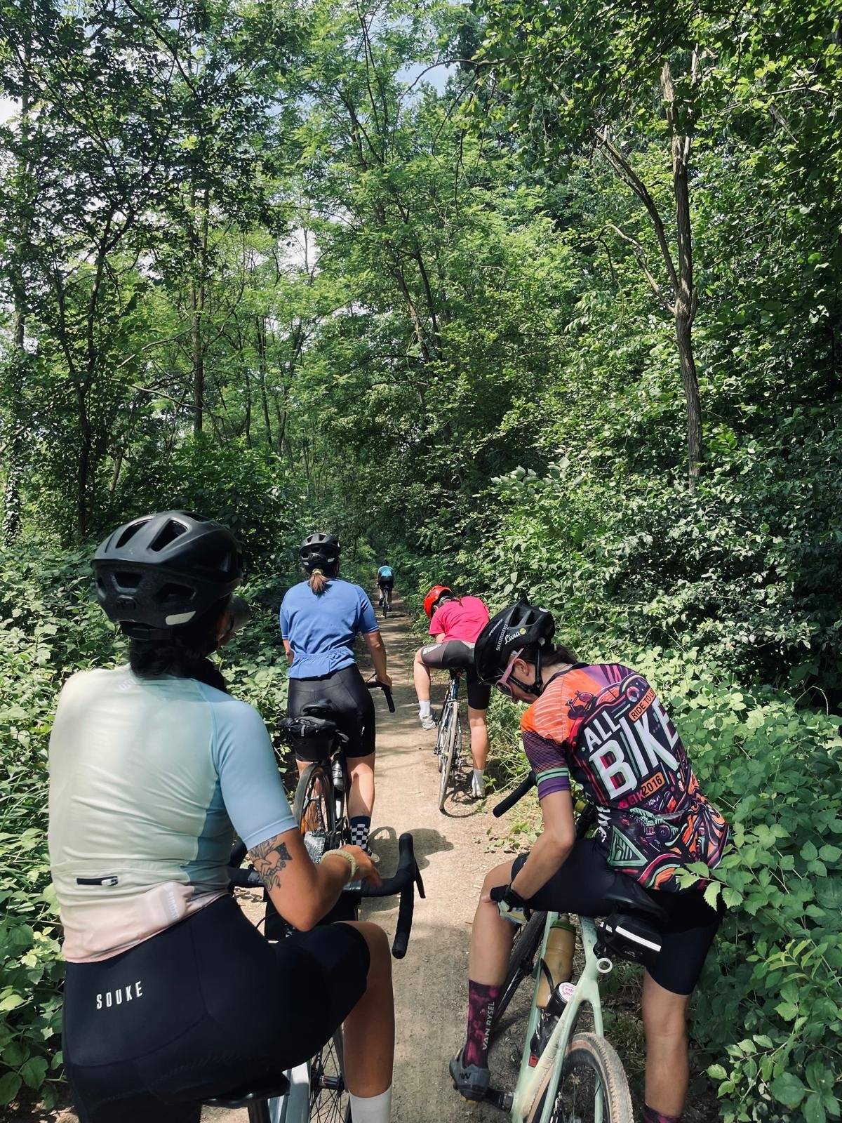 Four women on gravel bikes riding a narrow dirt path through dense green woodland