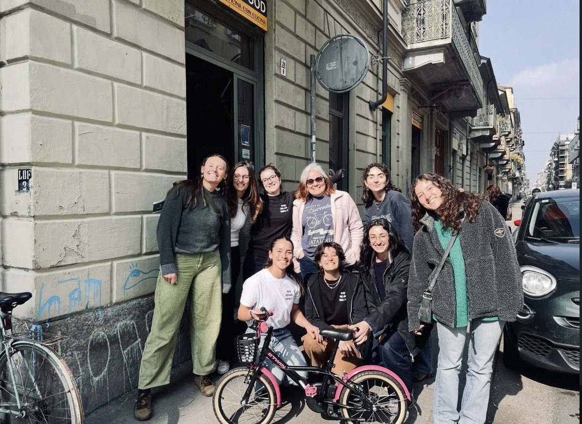 Ten women gathered on the pavement outside a Turin bike shop with a bicycle in front of them