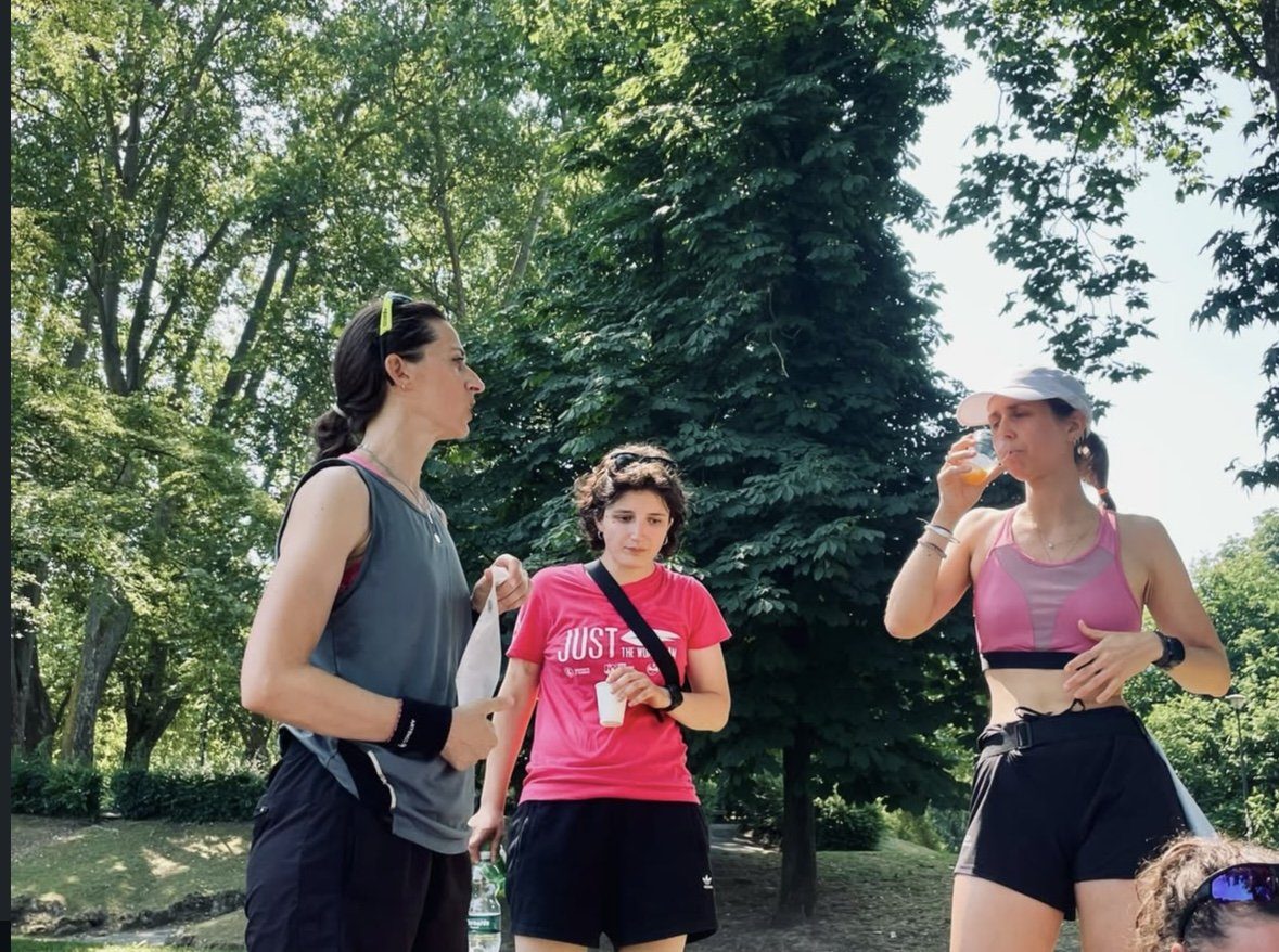 Three women standing in a sunny park in Turin after a group run, drinking juice and talking