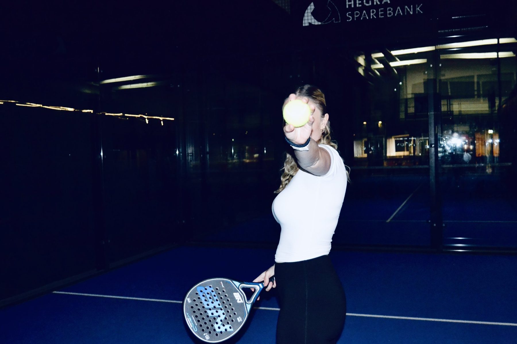 A woman holding a padel ball out towards the camera on a floodlit court at night