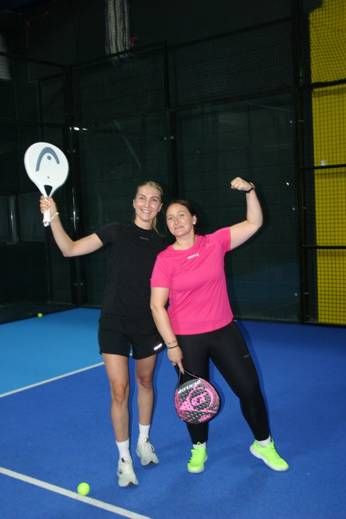 Two women posing with padel rackets on a blue court, one flexing her arm